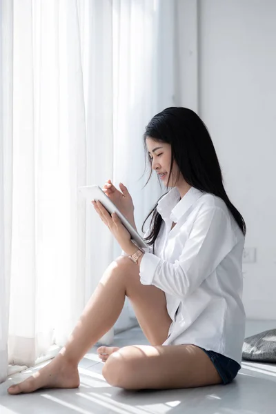 Piękna kobieta azjatyckich w białej koszuli przy użyciu tabletu w pobliżu okna w — Zdjęcie stockowe