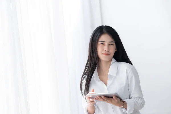 Hermosa mujer asiática en camisa blanca usando tableta cerca de ventana en —  Fotos de Stock