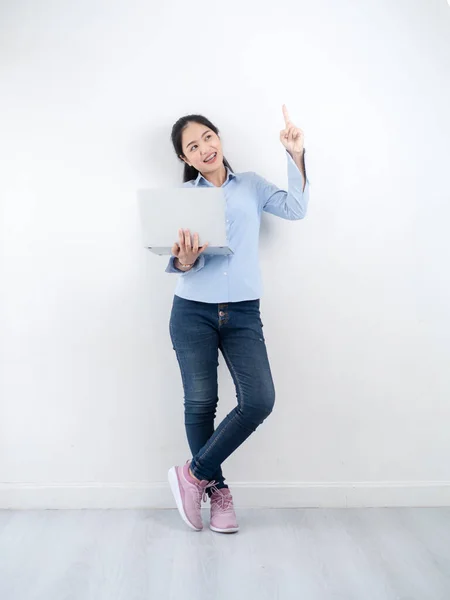 Happy Asian woman using laptop with white wall, pointing up. — Stockfoto