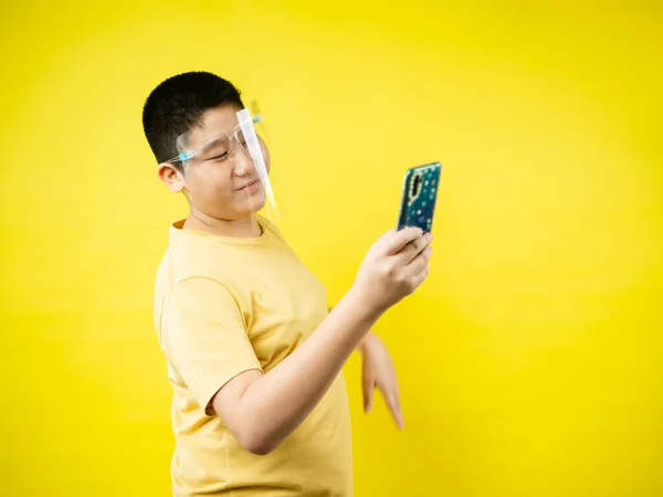 黄色の背景を持つスマートフォンで顔の盾と自撮りを身に着けているアジアの十代の少年 新しい通常の概念 — ストック写真