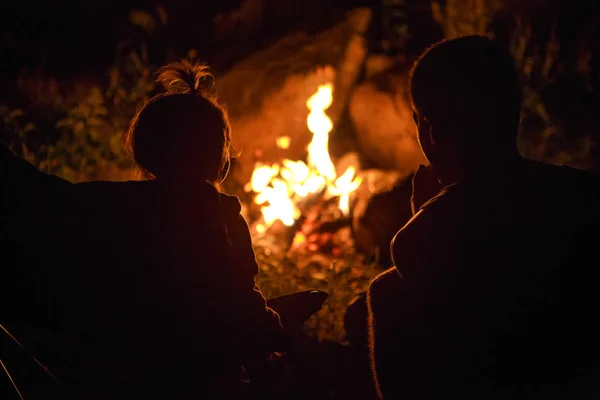 My daughter and father sit by the fire at night on a picnic in nature. Black silhouettes of a family against the fire