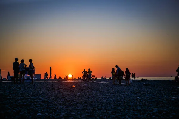 Black silhouettes of people on the beach in the sunset light of the orange sun. Pebble beach Batumi at sunset, tourists on the beach meet the sunset.