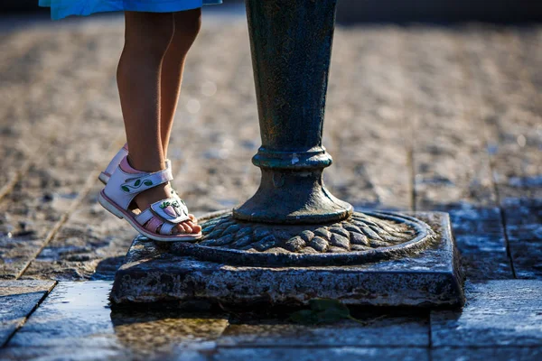 Baby feet in the sandals near the drinking fountain stood on the tips of his fingers. The child reaches up to the drinking fountain