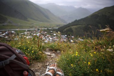 Dağlık bir bölgedeki bir köyün arka planında, turist beyaz sandaletleri içinde ayaklar. Köyün dağ manzarası, bir sırt çantasıyla patika boyunca yürüyor.