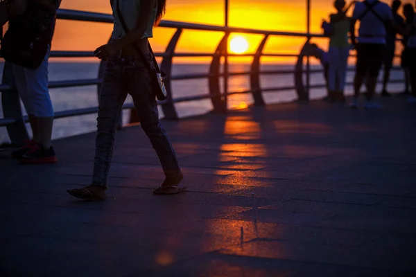 People walk along the promenade in the yellow and orange rays of the sun at sunset. Go on foot, ride a skateboard, silhouettes in the light of the sun and the shadows from the fence and railing. The sun shines through the railing