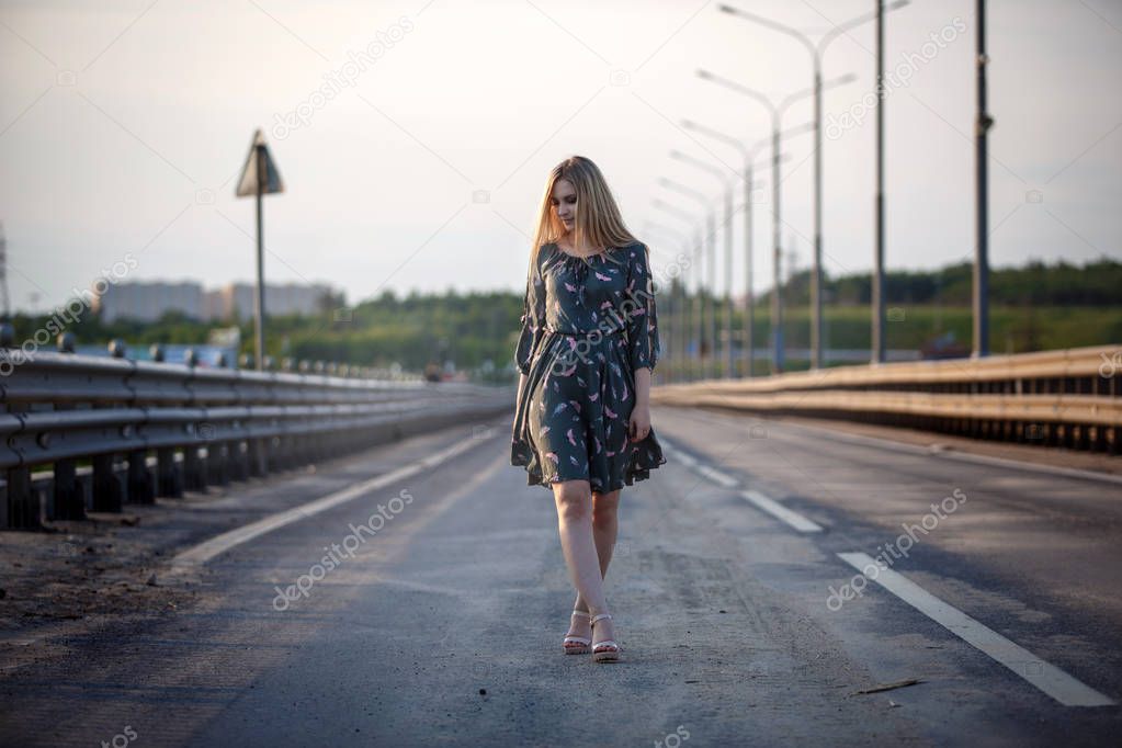 Young blonde girl on the road in the evening soft light of the sun. A girl in a green dress with long hair, the dress is blown by the wind.