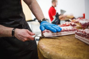 Et kesme ve doğrama işlemi. Elinde balta ve testere olan eldivenli bir adam eti özel bir tahta masada keser, kancalara asılı çiğ et parçaları.