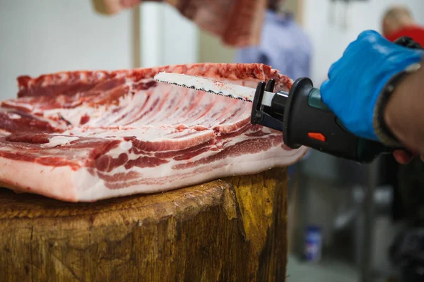 切肉的过程 一个戴着手套 戴着斧头和锯的男人把肉切在一张特殊的木桌上 一块生肉挂在钩子上 — 图库照片
