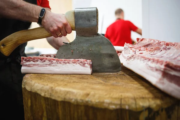 The process of cutting and chopping meat. A man in gloves with an axe and a saw cuts the meat on a special wooden table, pieces of raw meat hanging on hooks