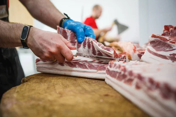 The process of cutting and chopping meat. A man in gloves with an axe and a saw cuts the meat on a special wooden table, pieces of raw meat hanging on hooks