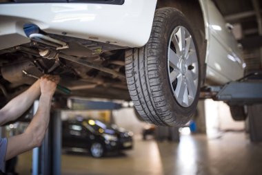 Servis istasyonunda araba tamiri. Direksiyonsuz asansörde arabayı tamir eden, lastiği tutan ve mekanik işleri tutan tulumda bir tamircinin elleri.