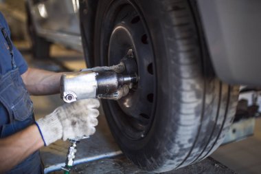 Servis istasyonunda araba tamiri. Direksiyonsuz asansörde arabayı tamir eden, lastiği tutan ve mekanik işleri tutan tulumda bir tamircinin elleri.