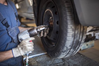 Servis istasyonunda araba tamiri. Direksiyonsuz asansörde arabayı tamir eden, lastiği tutan ve mekanik işleri tutan tulumda bir tamircinin elleri.