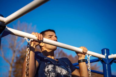 Kafkas görünümlü genç bir adam kendini yatay bir çubuğa çekiyor. Adam tek koluyla asılıyor. Sağlıklı yaşam tarzı, ekonomik açık hava sporları.