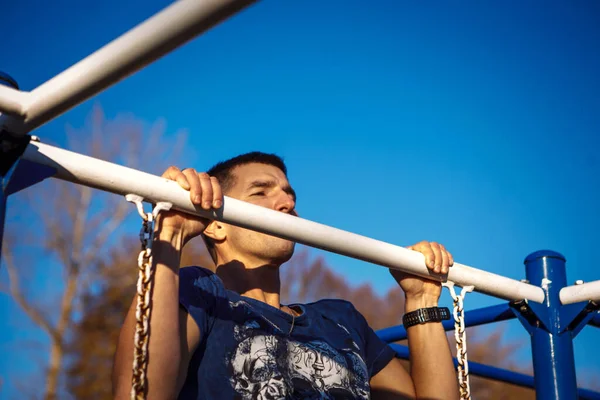 A young man of Caucasian appearance pulls himself up on a horizontal bar. The guy is hanging on one arm. Healthy lifestyle, affordable outdoor sports — Stock Image Young Man Caucasian Appearance Pulls Himself Horizontal Bar Guy Hanging Stock Image