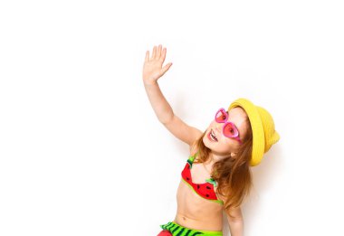 Plaj kıyafeti giymiş bir kız el sallıyor ve mutlu. Deniz tatilleri için çocuk aksesuarları: şapka, gül rengi kalp şeklinde bardaklar, karpuz desenli mayo. Güneş koruması, plaj modası, kızım seyahat etmeye hazır. Metin için beyaz arkaplan