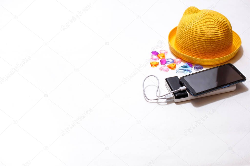 Smartphone on an external battery charge next to beach accessories: yellow straw hat, seashells, sunglasses. The concept of a beach holiday, always connected, full battery, portable charger. Mockup