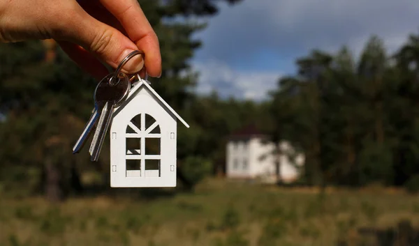 Wooden key chain and keys in hand on the background of an unfinished house. Dream of moving, the cottage is in a rural location and completion of the project. Space for text
