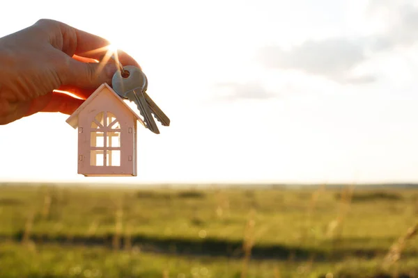 White wooden figure of a house and keys in hand against the background of the sky and field. Dream of your home, building a cottage in the countryside, plan and design, delivery of the project, moving to a new house.