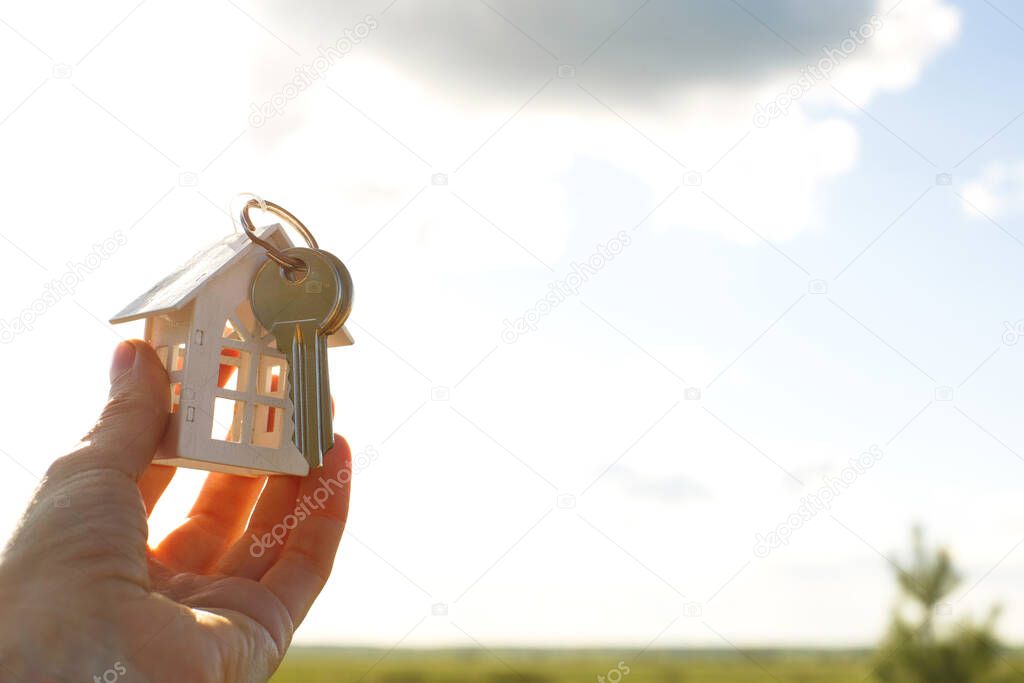 White wooden figure of a house and keys in hand against the background of the sky and field. Dream of your home, building a cottage in the countryside, plan and design, delivery of the project, moving to a new house.