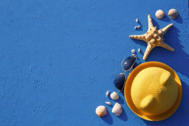 Deniz temalı plaj aksesuarları olan çerçeve: sarı hasır şapka, güneş gözlüğü, deniz yıldızı ve mavi arka planda deniz kabuğu. Tatil konsepti, deniz yolculuğu, ultraviyole koruma, yüzme. Uzayı kopyala Düz Yatay