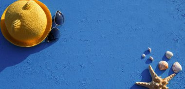 Deniz temalı plaj aksesuarları olan çerçeve: sarı hasır şapka, güneş gözlüğü, deniz yıldızı ve mavi arka planda deniz kabuğu. Tatil konsepti, deniz yolculuğu, ultraviyole koruma, yüzme. Uzayı kopyala Düz Yatay