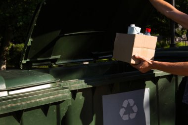 Ayrı atık toplama için konteynırların yanında elinde çöp olan bir karton kutu. Nüfusa ilişkin ekolojik farkındalık, çevreye özen, ayrılma: plastik, karton, ambalaj. 