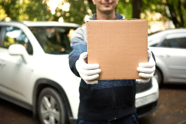 A cardboard box with space for text in the hands of a male courier in white gloves. Courier on the background of a white car. Contactless home delivery service for online orders. Mockup