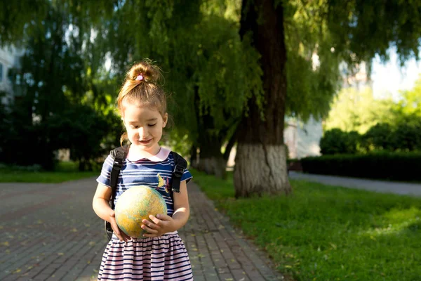 A girl from an elementary school of Caucasian appearance with a backpack on her shoulders holds a globe in her hands and carefully studies it. Education, return to school, September 1. Copy space