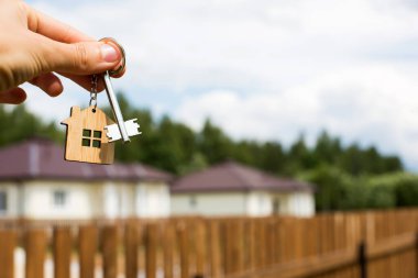 Bir ev ve anahtarın tahta kolyesi. Çitlerin ve kulübenin arka planı. Ev, bina, tasarım, projenin teslimi, yeni bir eve taşınma, ipotek, kira ve emlak satın alma hayali. Boşluğu kopyala