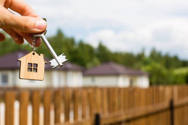 Bir ev ve anahtarın tahta kolyesi. Çitlerin ve kulübenin arka planı. Ev, bina, tasarım, projenin teslimi, yeni bir eve taşınma, ipotek, kira ve emlak satın alma hayali. Boşluğu kopyala