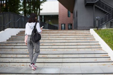 Elinde siyah bir sırt çantası ve elinde not defteri olan bir kız öğrenci üniversitenin önündeki basamaklarda duruyor. Yüksek öğrenim, okul yılının başlangıcı, okula dönüş. Kopyalama alanı