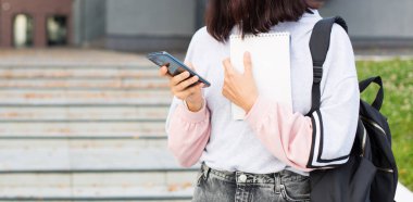 Siyah sırt çantalı bir kız öğrenci üniversitenin eşiğinde akıllı telefon ve not defteri tutuyor. Yüksek öğrenim, okul yılının başlangıcı, okula dönmek, aletler. Kopyalama alanı