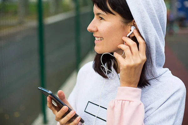 A Caucasian woman uses a smartphone via wired headphones with a headset. Phone in hand, white wire in ear, Enjoy label on hoodie. Listen to music via the gadget, sports in the app. Space for text