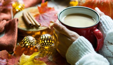 Düşmüş akçaağaç yapraklı sıcak kakaolu kırmızı bardak, yuvarlak lambalar, çelenkler, tarçın çubukları, sıcak battaniye. Sonbahar atmosferi, ılık kahve, beyaz kazaklı kadın elleri, rahatlık ve rahatlık.
