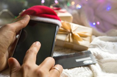 Noel Baba şapkalı akıllı telefon, Noel süslemelerinin arka planında banka kartı hediye kutuları, çelenkler. Yılbaşı alışverişi, uygulama üzerinden hediyeler almak, online mağaza. İnternetten alışveriş