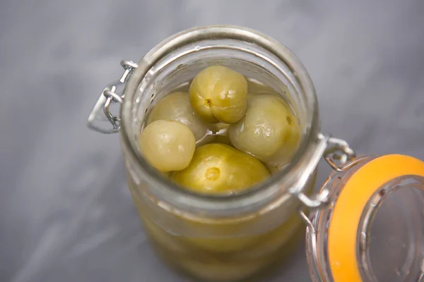 Pickled green small tomatoes in a glass jar with a lid on a gray background. Salted cherries, home-made, olives in brine.