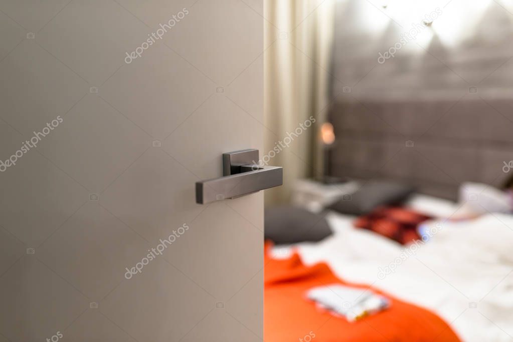 Ajar door to a modern bedroom with a light, visible blurred bed, bedding and orange pillows.