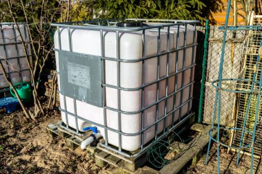 Large industrial liquid tank with a capacity of 1000 liters, 265 gallons with a metal protective grille. Placed in a home garden as a rainwater tank. clipart