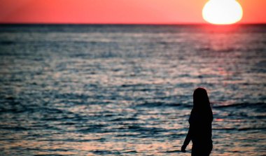 Deniz kıyısında duran ve güzel bir gün batımına bakan genç bir kadının silueti.
