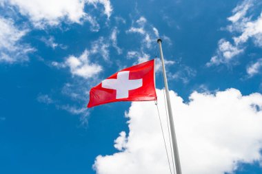 Sola doğru rüzgarda sallanan, İsviçre bayrağı direğe asılı, beyaz bulutlar ile mavi gökyüzü nün arka planında.