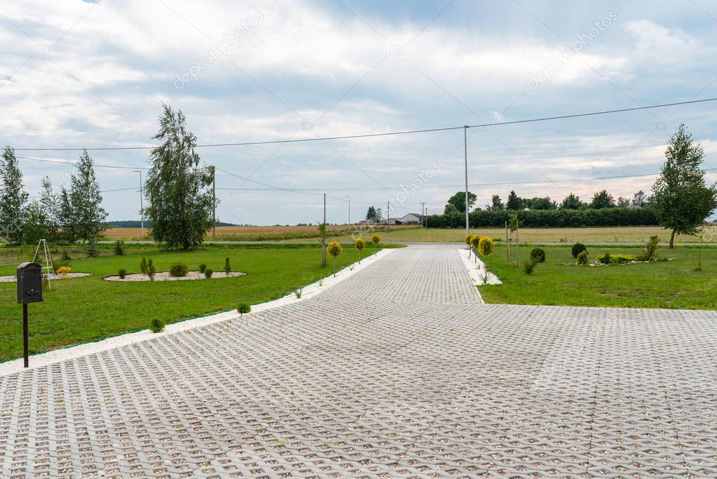 Entry for cars into the property made of openwork concrete pavement filled with colorful pebbles, around is green grass and small trees.