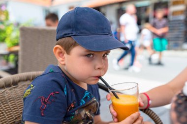 Bir restoranda bir hasır sandalyede oturan bir lacivert beyzbol şapkası beş yıllık sevimli çocuk ve bir içme saman portakal suyu içiyor.