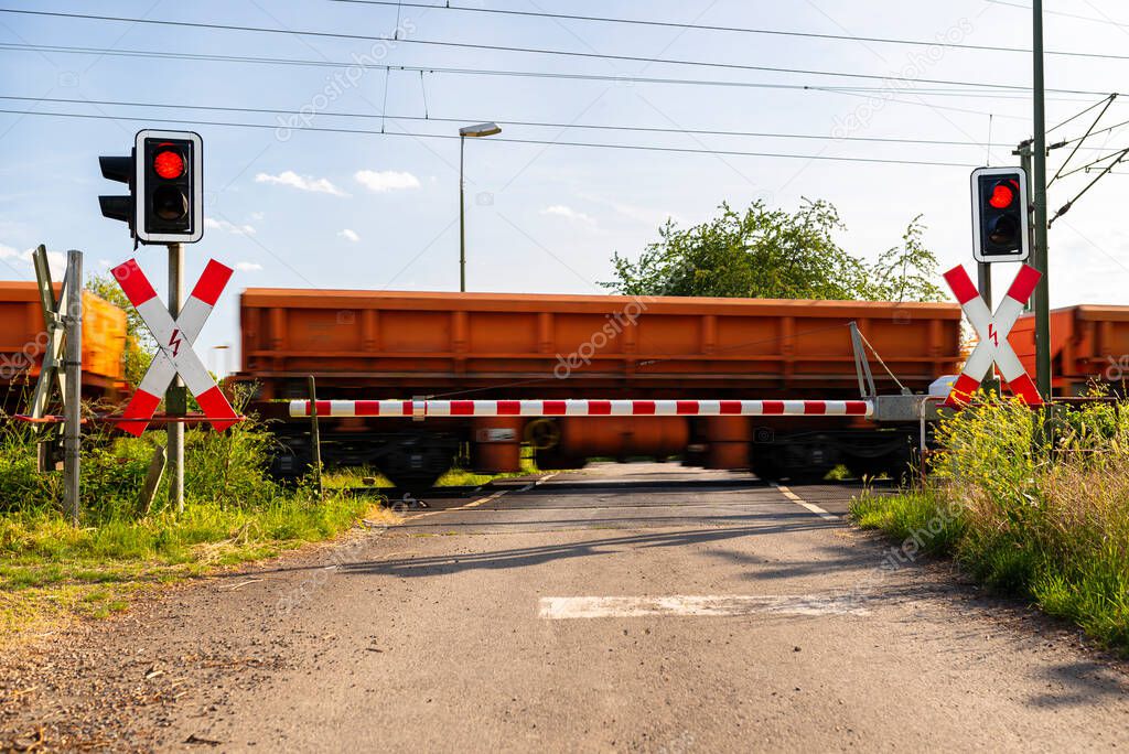 Closed barrier at the railroad crossing with red warning lights on, visible blurred red wagons in motion. 