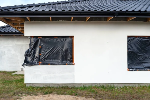 A newly applied layer of white silicone plaster on the wall of the house, the corner window is protected with black foil.