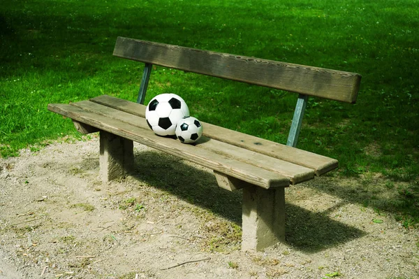 two soccer balls, one big, one small are laying together on a park bench