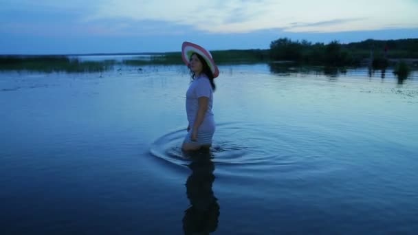 Una giovane ragazza in pantaloncini e un cappello passeggia lungo il lago e ammira il tramonto . . — Video Stock