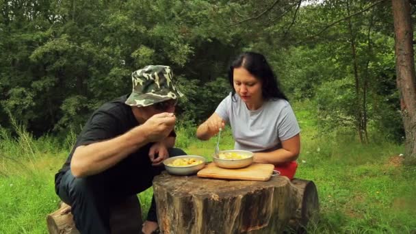 Um homem e uma mulher estão sentados à beira de uma floresta em árvores caídas e comendo salada de frutas. . — Vídeo de Stock