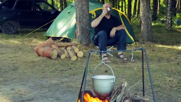 Muž sedí na stan, pije čaj a sleduje tvrďák visí nad ohněm. — Stock video