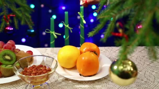 Fruits and nuts with honey are on the table with a decorated Christmas tree ball and the lights are blurry bokeh. In the background, the woman lights New Year candles.